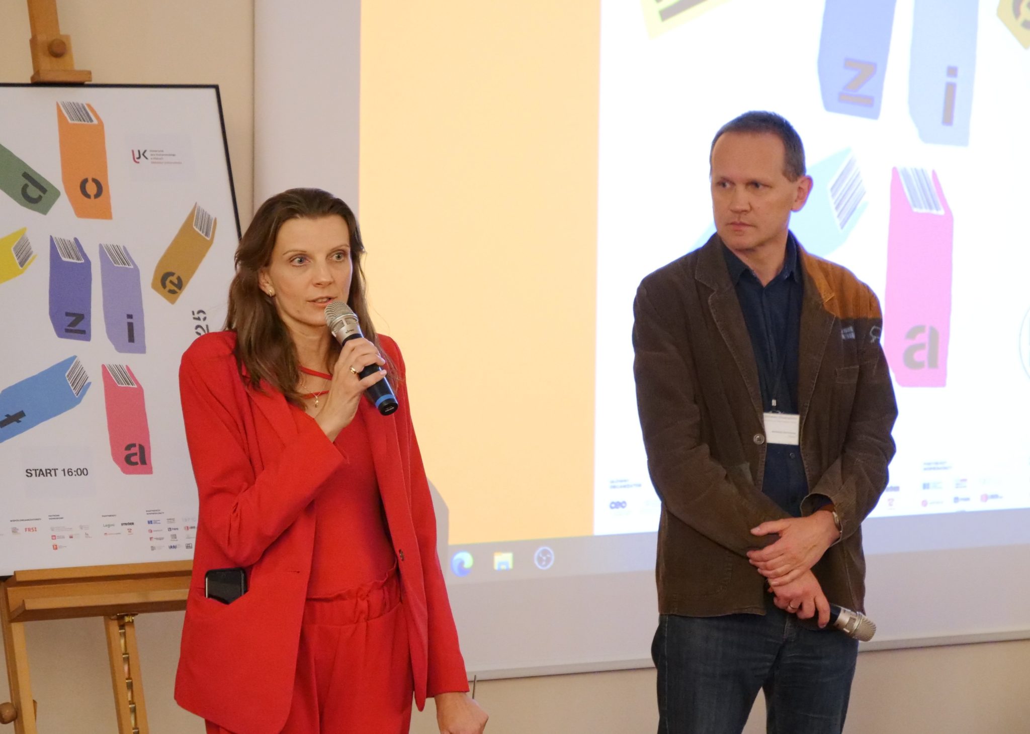 Kobieta w czerwonym garniturze mówi do mikrofonu, obok stoi mężczyzna z mikrofonem w dłoni. Oboje znajdują się na tle ekranu z kolorową grafiką przedstawiającą książki i napisem „Noc Bibliotek 2025"