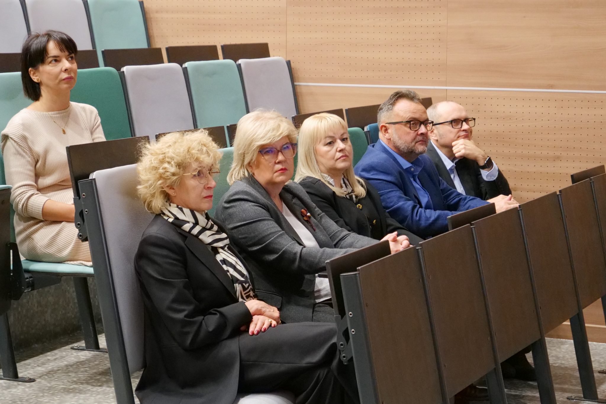 Pięć osób – trzy kobiety i dwaj mężczyźni – siedzi w pierwszym rzędzie auli. Wszyscy ubrani są w formalne lub półformalne stroje i z uwagą patrzą w lewo (z perspektywy widza), słuchając wykładu. Na pierwszym planie kobieta w czarnym garniturze i szaliku.