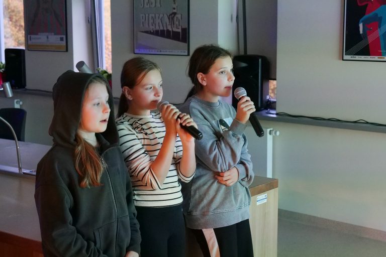 Trzy dziewczynki trzymają w dłoniach mikrofony i śpiewają.