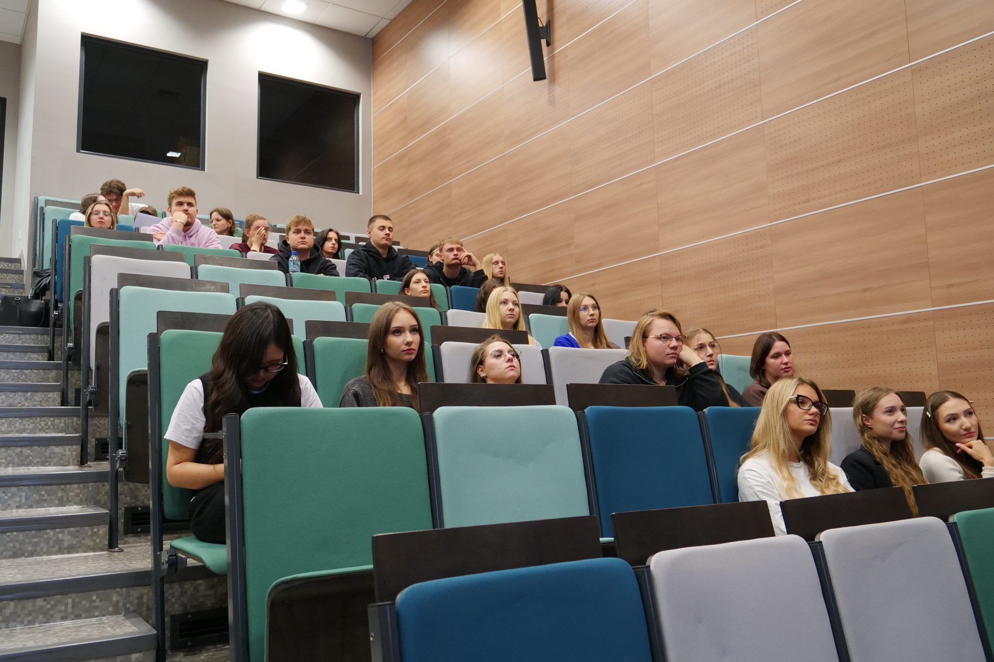 Szerokie ujęcie auli wykładowej z rzędami zielonych i niebieskich krzeseł. Kilka rzędów jest częściowo zapełnionych przez studentów, którzy siedzą i patrzą w jednym kierunku, prawdopodobnie na prelegenta. Ściany auli są wyłożone jasną, drewnianą boazerią.