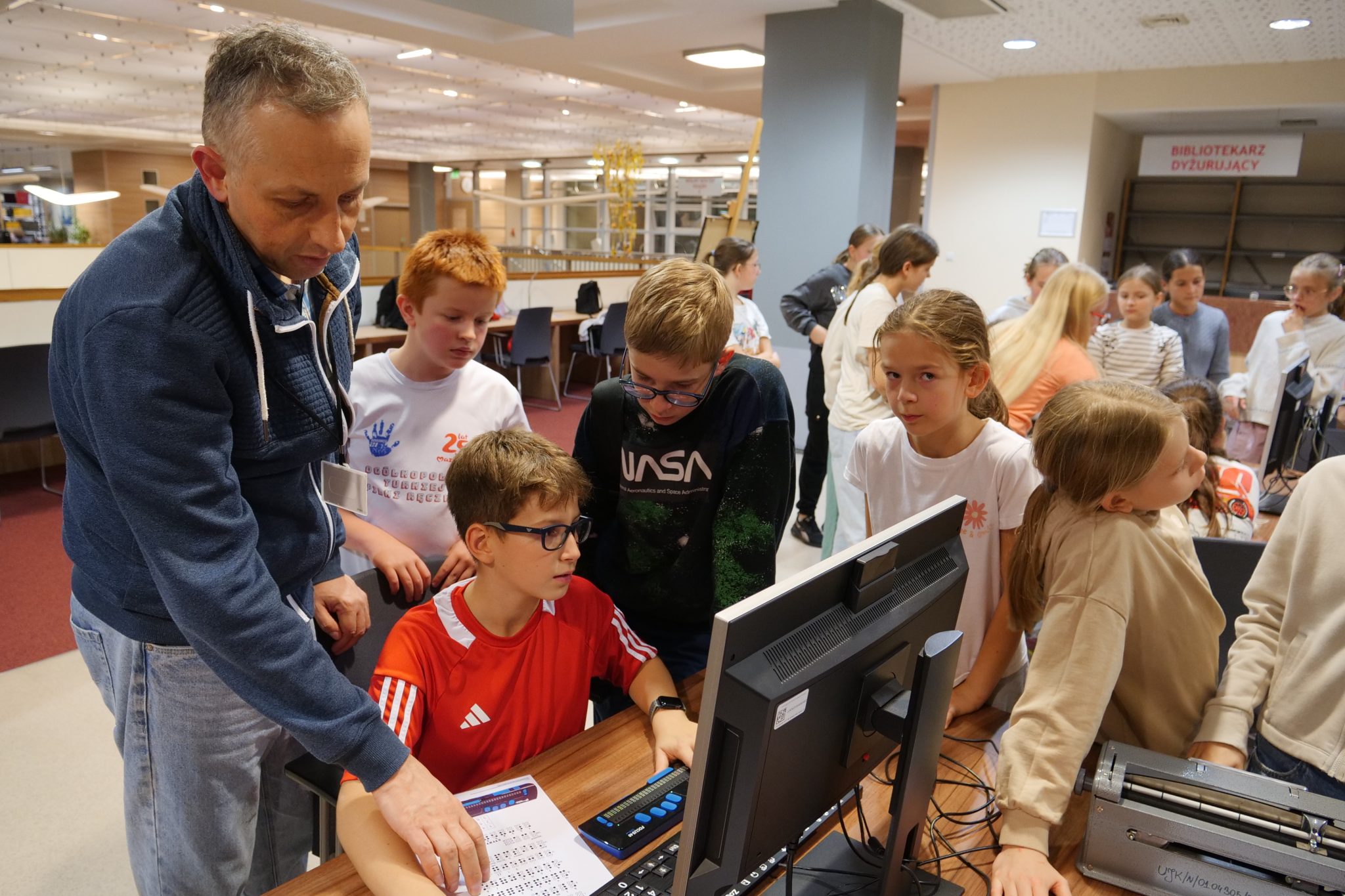 Grupa dzieci siedzi wokół komputera. Obok nich stoi wysoki mężczyzna w niebieskiej bluzie. Wskazuje dłonią na kartkę papieru, na której znajduje się alfabet Braille'a.