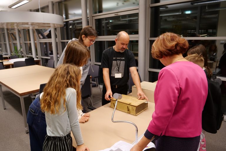 Mężczyzna ubrany w czarną koszulkę stoi przy stole, na którym leży stara, gruba książka. Dookoła stołu stoi kilkoro dzieci.