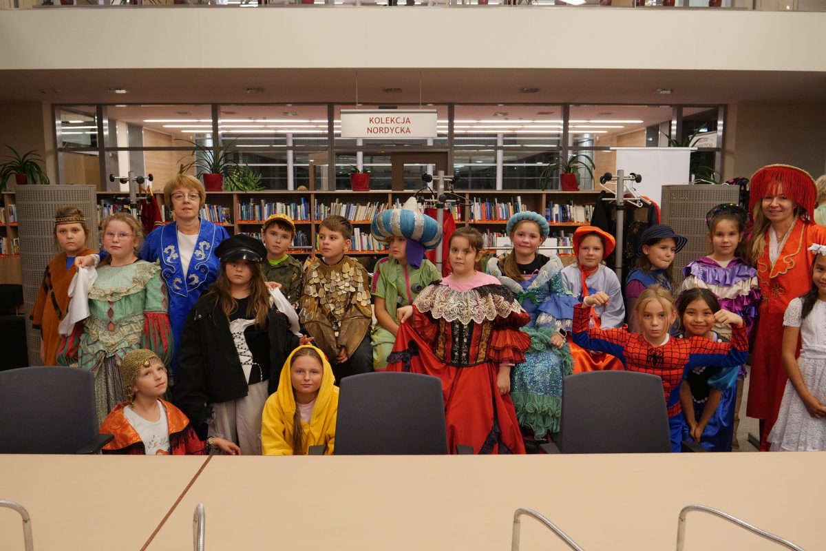 Grupa dzieci i dwie dorosłe kobiety pozują w bibliotece. Wszyscy mają na sobie kolorowe kostiumy: historyczne stroje, bajkowe przebrania, m.in. księżniczki, orientalni książęta, Spiderman. W tle regały z książkami i napis „Kolekcja Nordycka”.