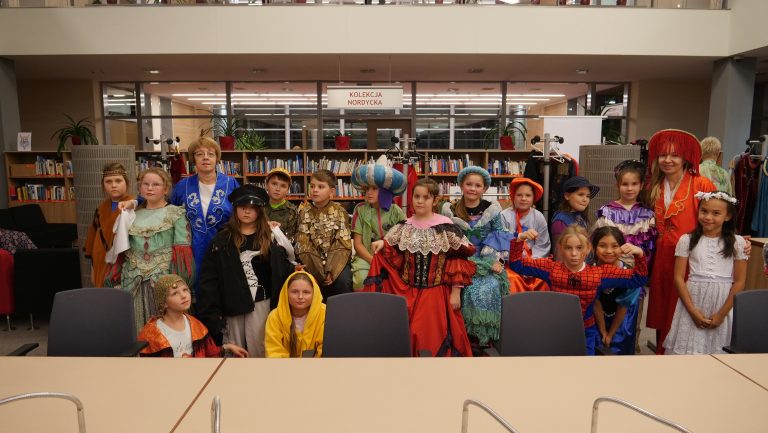Grupa dzieci i dwie dorosłe kobiety pozują w bibliotece. Wszyscy mają na sobie kolorowe kostiumy: historyczne stroje, bajkowe przebrania, m.in. księżniczki, orientalni książęta, Spiderman. W tle regały z książkami i napis „Kolekcja Nordycka”.