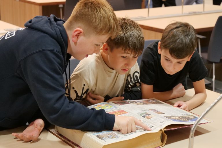 Trzech chłopców uważnie przegląda dużą książkę lub album na stole w czytelni. Dwóch z nich pochyla się, czytając, a jeden palcem wskazuje na fragment tekstu. W tle widać puste stanowiska biblioteczne z lampkami.