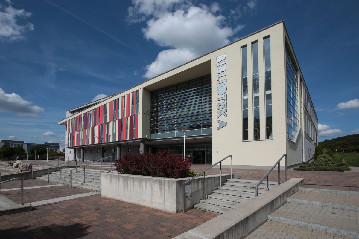 Nowoczesny budynek biblioteki z napisem „BIBLIOTEKA” na jasnej elewacji. Przód budynku ozdobiony kolorowymi panelami i dużymi oknami. Przed wejściem schody, podjazd i roślinność. Pogodny dzień z błękitnym niebem i chmurami.
