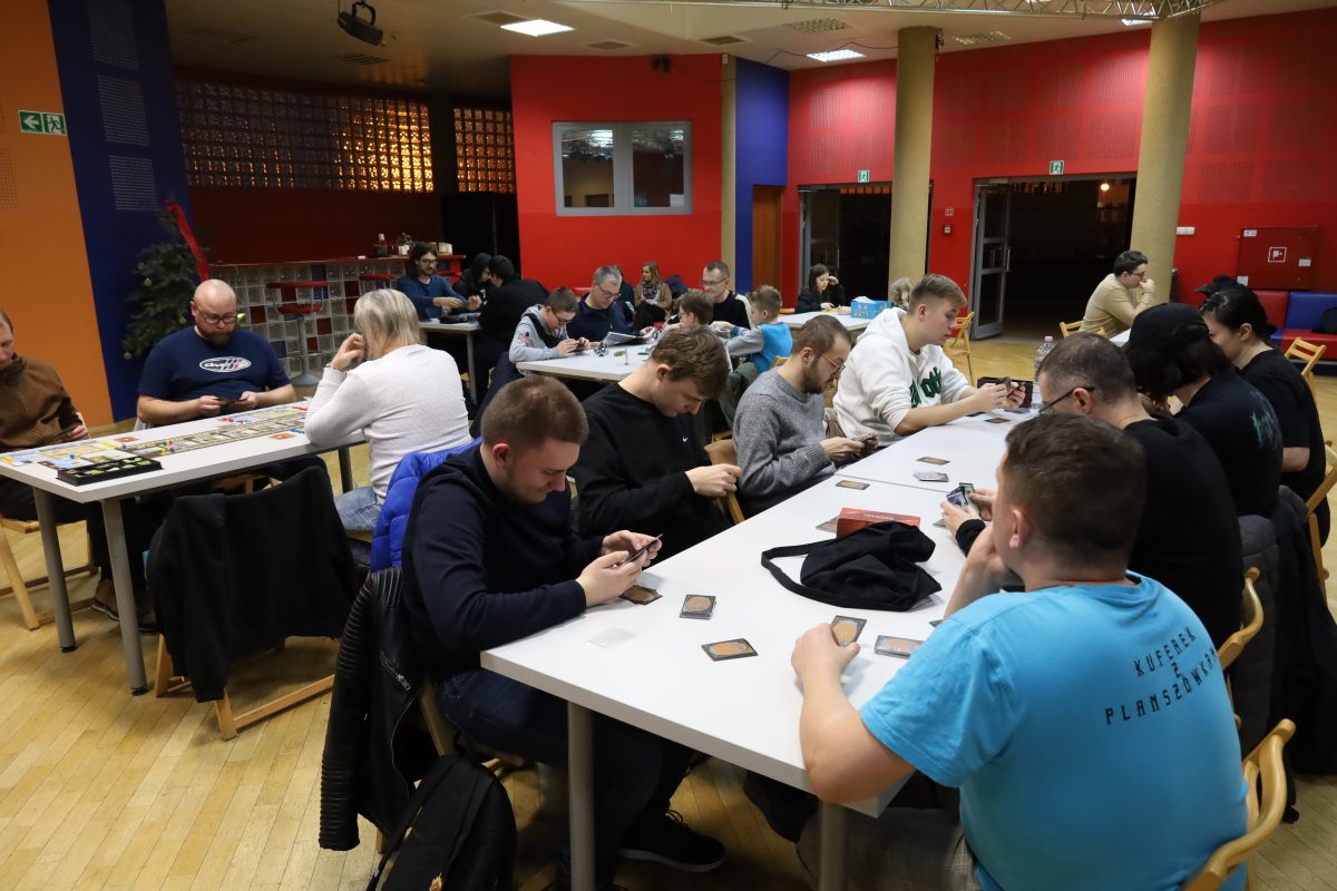 Na zdjęciu widać salę z kilkoma stołami, przy których siedzi wiele osób grających w gry planszowe. Wszyscy są skupieni na kartach i pionkach trzymanych w dłoniach. Pomieszczenie jest kolorowe, z czerwonymi, niebieskimi i pomarańczowymi ścianami oraz jasnym oświetleniem.