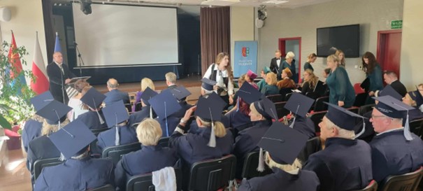 Na zdjęciu widać salę wykładową, prowadzącego wykład mężczyznę. Na sali widoczni są Kobiety i Mężczyźni, którzy słuchają wykładu i patrzą na wyświetlaną prezentację.