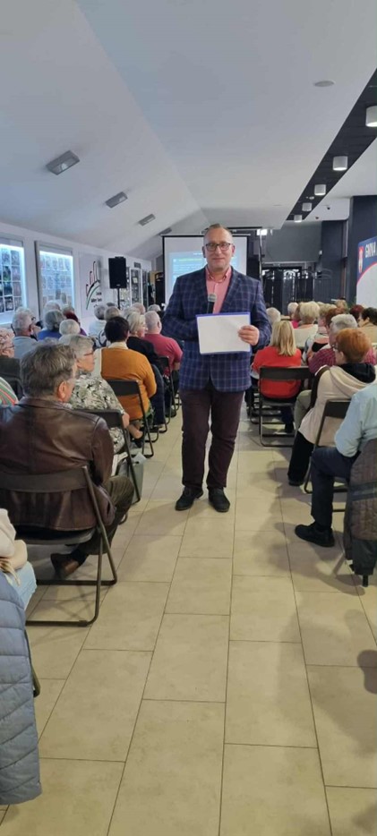 Na zdjęciu widać salę wykładową, prowadzącego wykład mężczyznę. Na sali widoczni są Kobiety i Mężczyźni, którzy słuchają wykładu i patrzą na wyświetlaną prezentację.