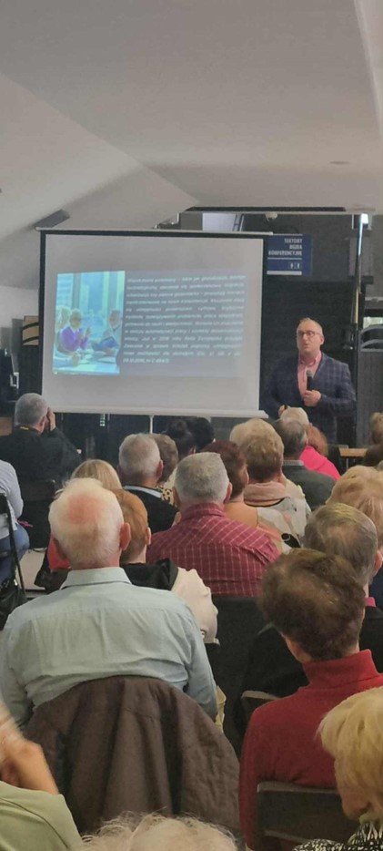 Na zdjęciu widać salę wykładową. Na sali widoczni są Kobiety i Mężczyźni, którzy słuchają wykładu i patrzą na wyświetlaną prezentację.