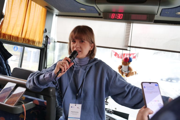 Młoda kobieta w niebieskiej bluzie z identyfikatorem Erasmus+ mówi do mikrofonu w autokarze, prowadząc wycieczkę. W tle widać zawieszkę z napisem „Polska” i pluszową maskotkę. Na siedzeniu obok znajdują się dwa telefony komórkowe.