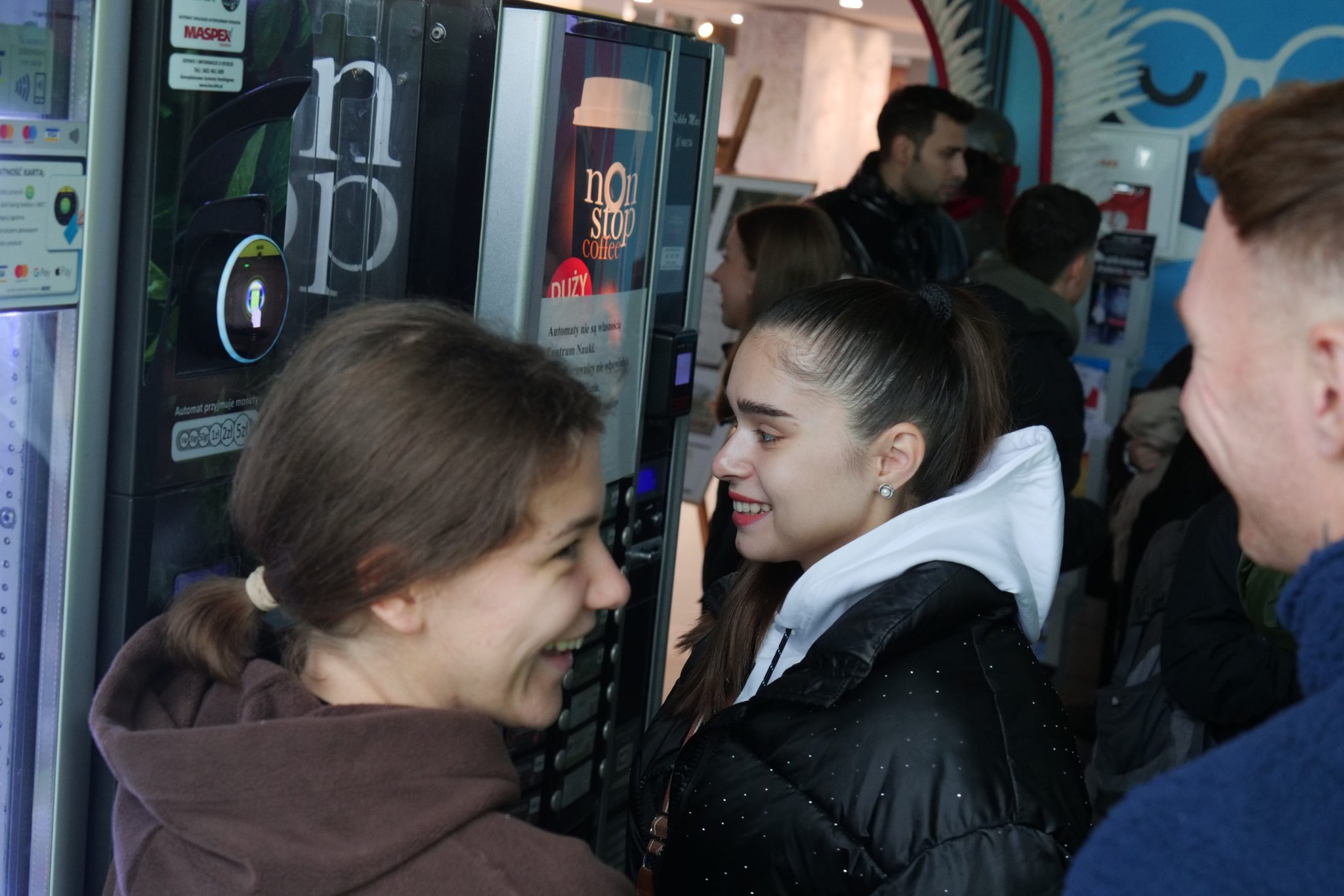 Grupa młodych osób, w tym dwie uśmiechnięte dziewczyny, stoi przy automacie z kawą. W tle widać innych studentów czekających w kolejce. Atmosfera jest luźna i towarzyska, a kolorowe otoczenie sugeruje, że znajdują się w przestrzeni edukacyjnej lub muzealnej.
