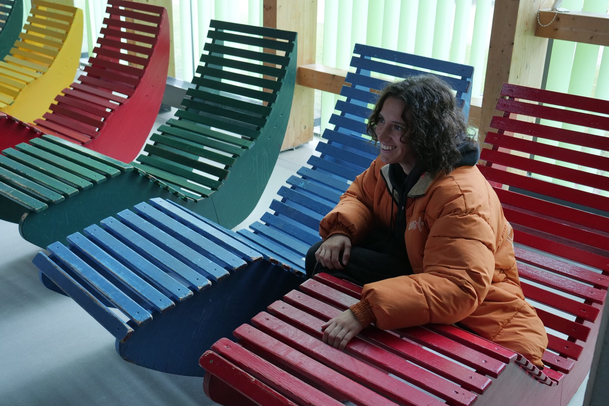 Uśmiechnięta młoda kobieta w pomarańczowej kurtce siedzi na kolorowym, designerskim leżaku wykonanym z drewnianych listew. Wokół niej znajdują się inne leżaki w intensywnych kolorach: czerwonym, niebieskim, zielonym i żółtym. Pomieszczenie jest jasne, z dużymi oknami i pionowymi żaluzjami.