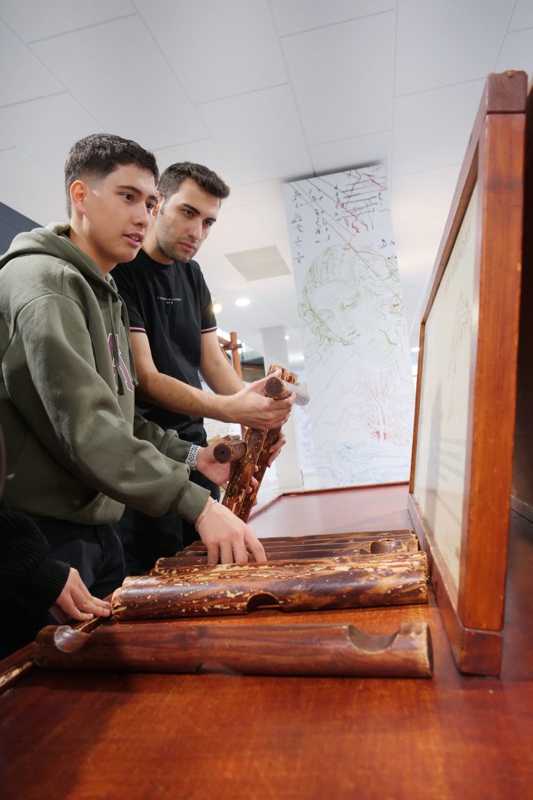 Dwaj młodzi mężczyźni w muzeum, stoją przy drewnianym eksponacie. Mężczyzna po lewej w zielonej bluzie z kapturem i mężczyzna po prawej w czarnej koszulce manipulują dużymi, rzeźbionymi drewnianymi elementami przypominającymi rury lub cylindry, które są ułożone na drewnianym blacie. Po prawej widoczna jest część drewnianej ramy. W tle widać białe ściany z rysunkami i tekstem w kolorze czerwonym.