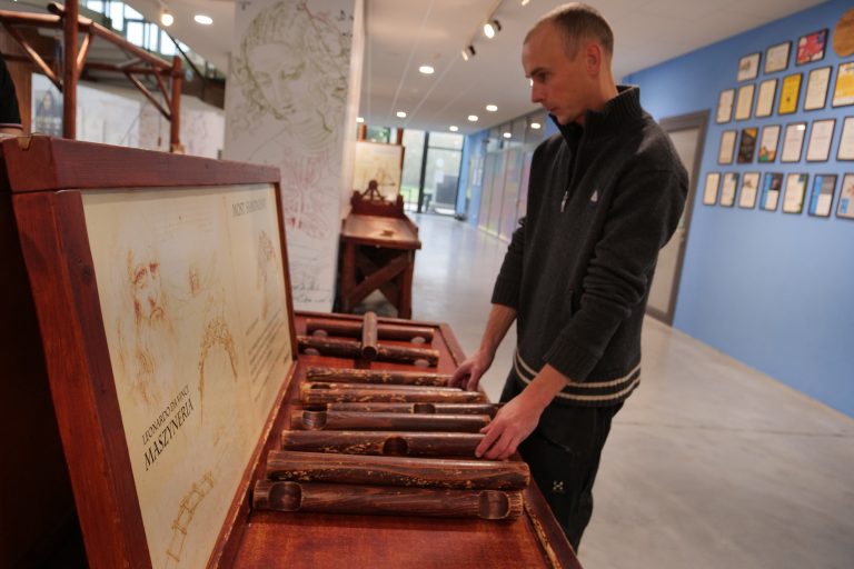 Mężczyzna ubrany w ciemnoszary sweter na suwak, stoi przy tym samym drewnianym eksponacie. Pochyla się, dotykając i układając rzeźbione, drewniane elementy. Po lewej stronie eksponatu znajduje się uchylona drewniana pokrywa z grafiką i tekstem w języku polskim, w tym słowami "LEONARDO DA VINCI", "PROJEKTY", i "MASZYNERIA". W tle widoczne jest jasne wnętrze z niebieską ścianą, na której wisi wiele oprawionych certyfikatów lub zdjęć, oraz inne eksponaty w dalszej części pomieszczenia.