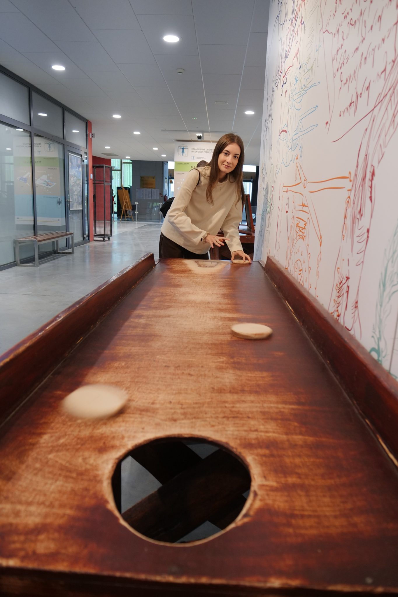 Młoda kobieta w beżowej bluzie stoi przy długim, drewnianym stole do gry, prawdopodobnie w shuffleboard, popychając drewniany krążek.