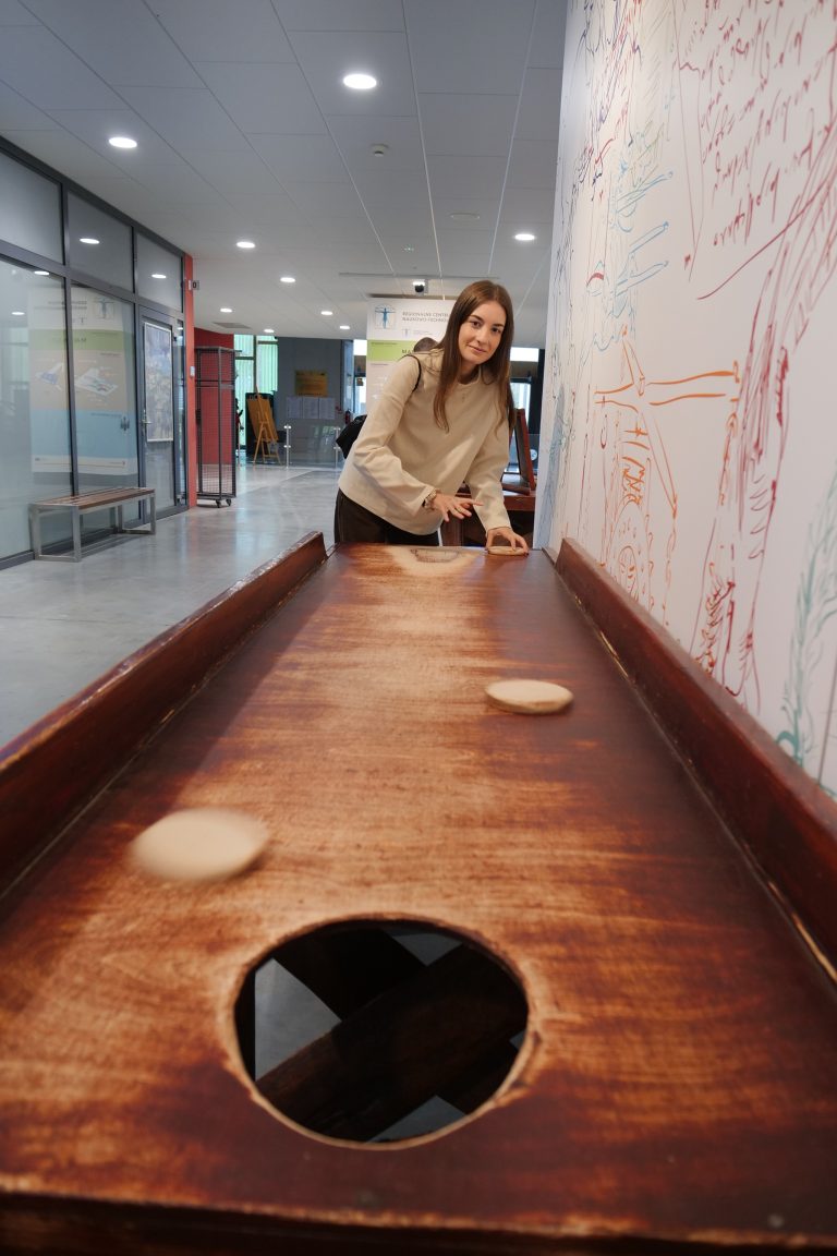 Młoda kobieta w beżowej bluzie stoi przy długim, drewnianym stole do gry, prawdopodobnie w shuffleboard, popychając drewniany krążek.