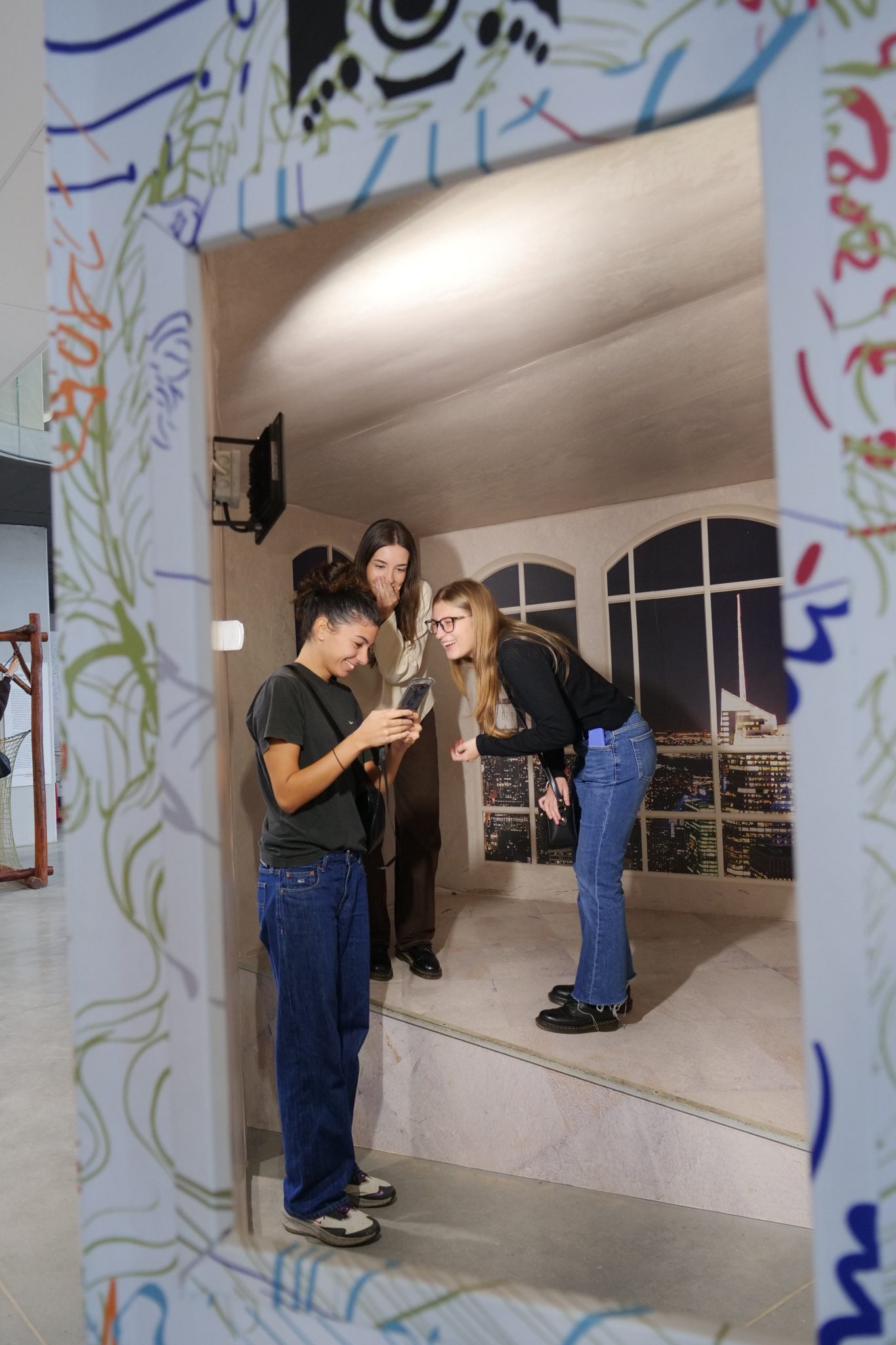 Trzy młode kobiety oglądają coś na telefonie komórkowym, stojąc w specjalnym pomieszczeniu o zniekształconej perspektywie, przypominającym pokój Eamesa, co sugeruje, że jest to iluzja optyczna.