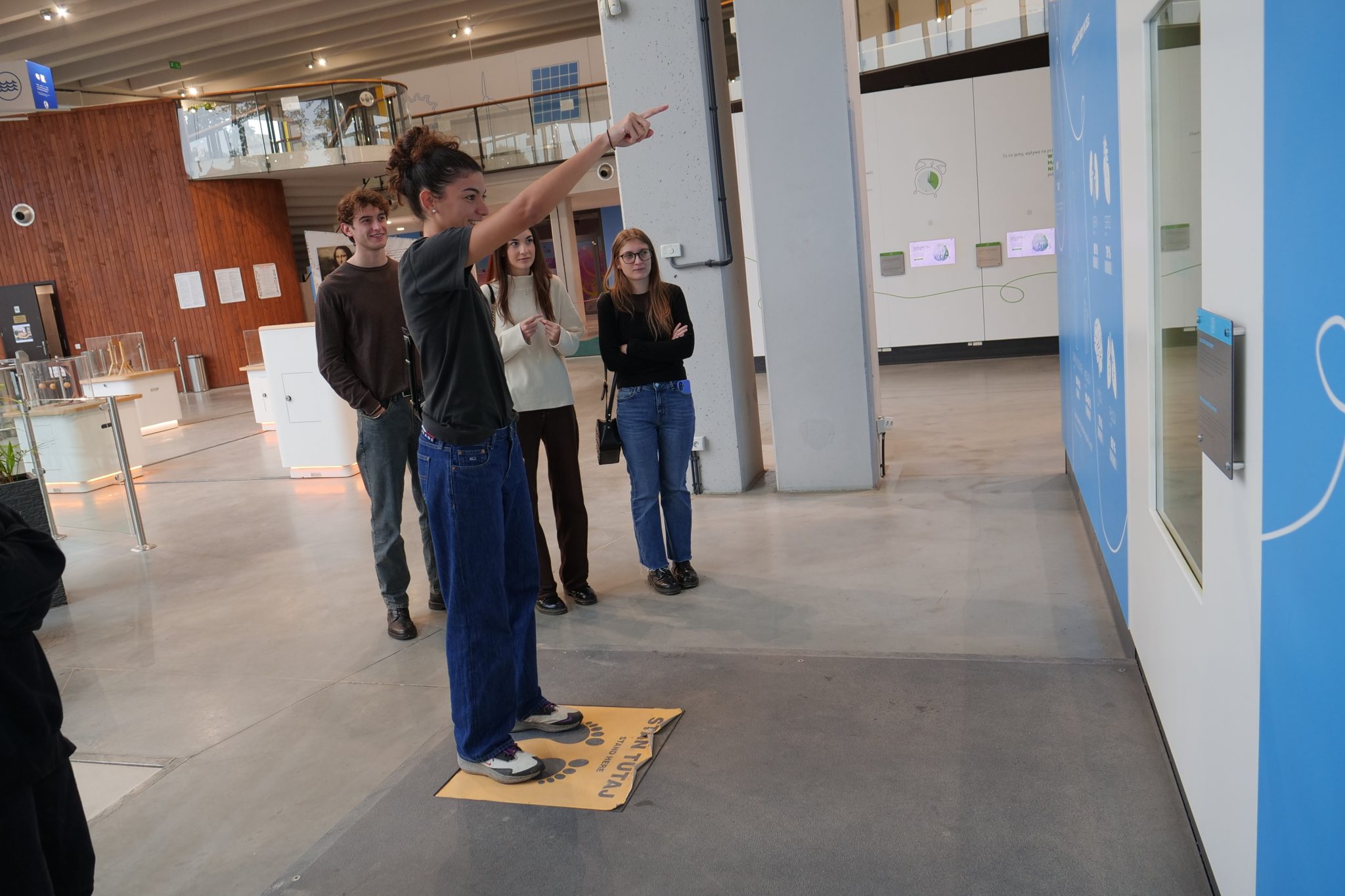 Młoda kobieta stojąca na żółtej macie, prawdopodobnie będącej częścią interaktywnego eksponatu, wskazuje palcem w prawo, podczas gdy troje innych młodych ludzi stoi za nią.