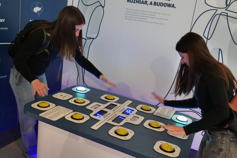 Dwie kobiety z długimi ciemnymi włosami wchodzą w interakcję z niebieskim, cyfrowym eksponatem w muzeum nauki, który zawiera żółte przyciski i podświetlane pola.