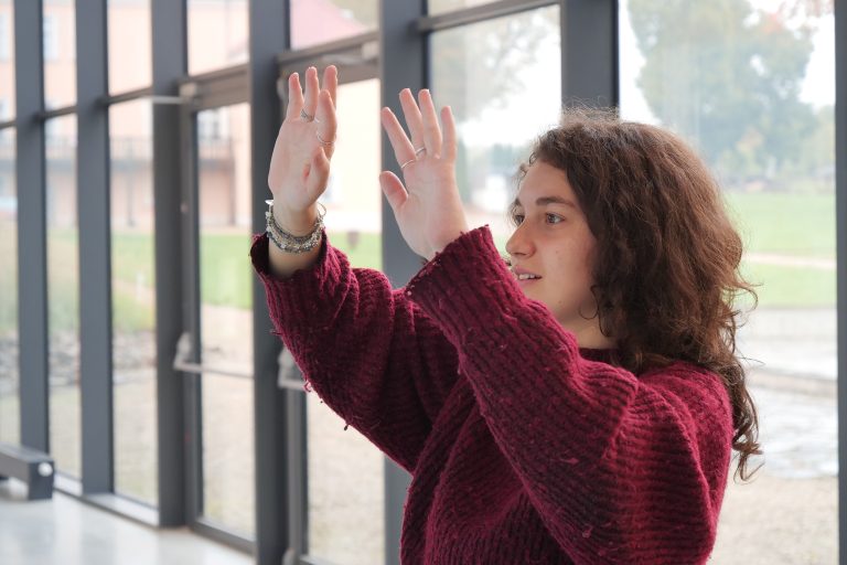 Młoda kobieta z kręconymi, ciemnymi włosami i w bordowym swetrze unosi ręce przed dużymi przeszklonymi drzwiami, patrząc z zaciekawieniem.