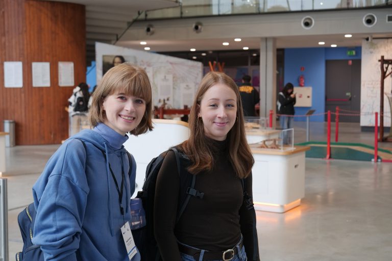 Dwie młode kobiety, uśmiechnięte, pozują razem do zdjęcia w jasnej, otwartej przestrzeni.