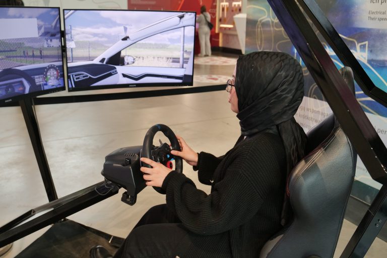 Młoda kobieta w ciemnym nakryciu głowy i okularach siedzi w symulatorze jazdy, trzymając kierownicę.
