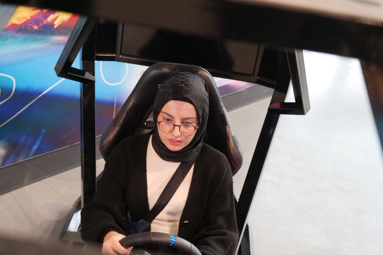 Młoda kobieta, ubrana na czarno, w ciemnym nakryciu głowy, siedzi w symulatorze jazdy samochodem, wpatrując się w ekran wyświetlający wirtualną trasę.