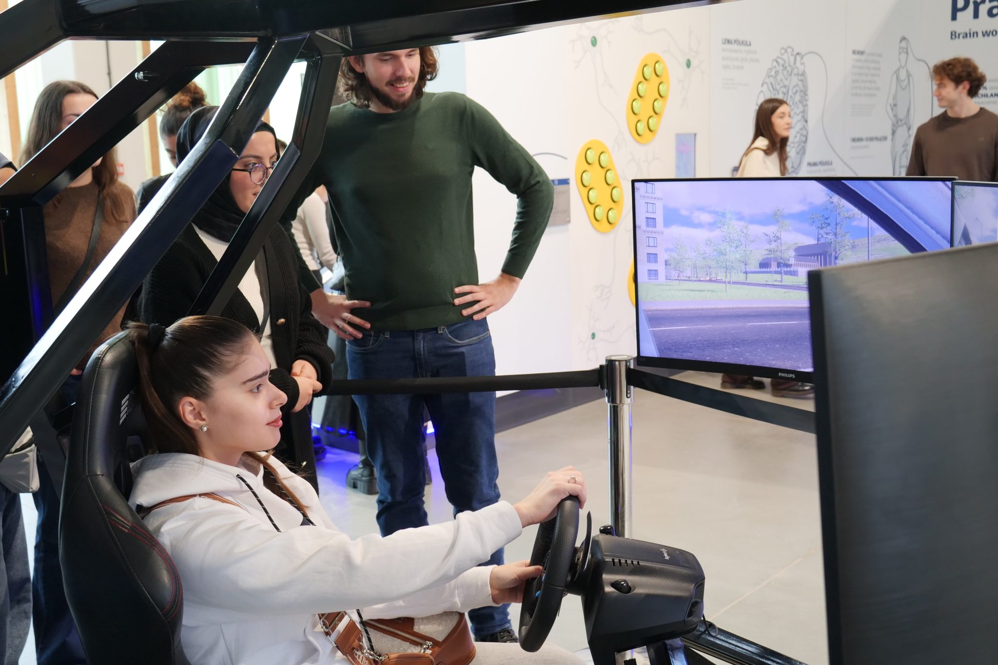 Młoda kobieta w białej bluzie z kapturem siedzi w symulatorze jazdy i trzyma kierownicę, skupiona na ekranie przedstawiającym wirtualne miasto.