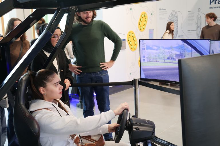 Młoda kobieta w białej bluzie z kapturem siedzi w symulatorze jazdy i trzyma kierownicę, skupiona na ekranie przedstawiającym wirtualne miasto.