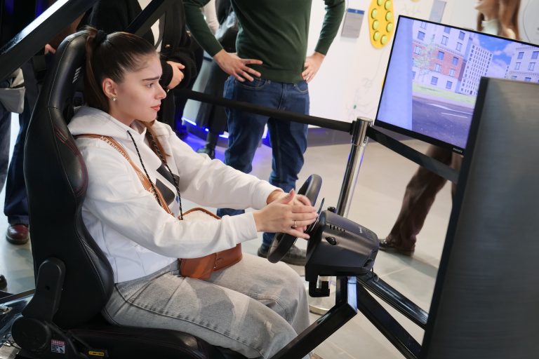 Młoda kobieta w białej bluzie z kapturem siedzi w symulatorze jazdy i trzyma kierownicę, skupiona na ekranie przedstawiającym wirtualne miasto.