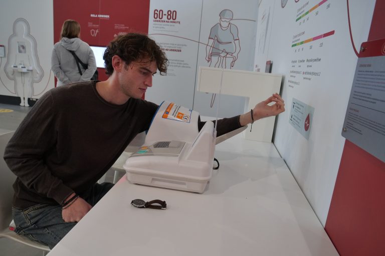 Młody mężczyzna w brązowym swetrze używa automatu do pomiaru ciśnienia krwi, który jest częścią eksponatu na białym stole w muzeum.