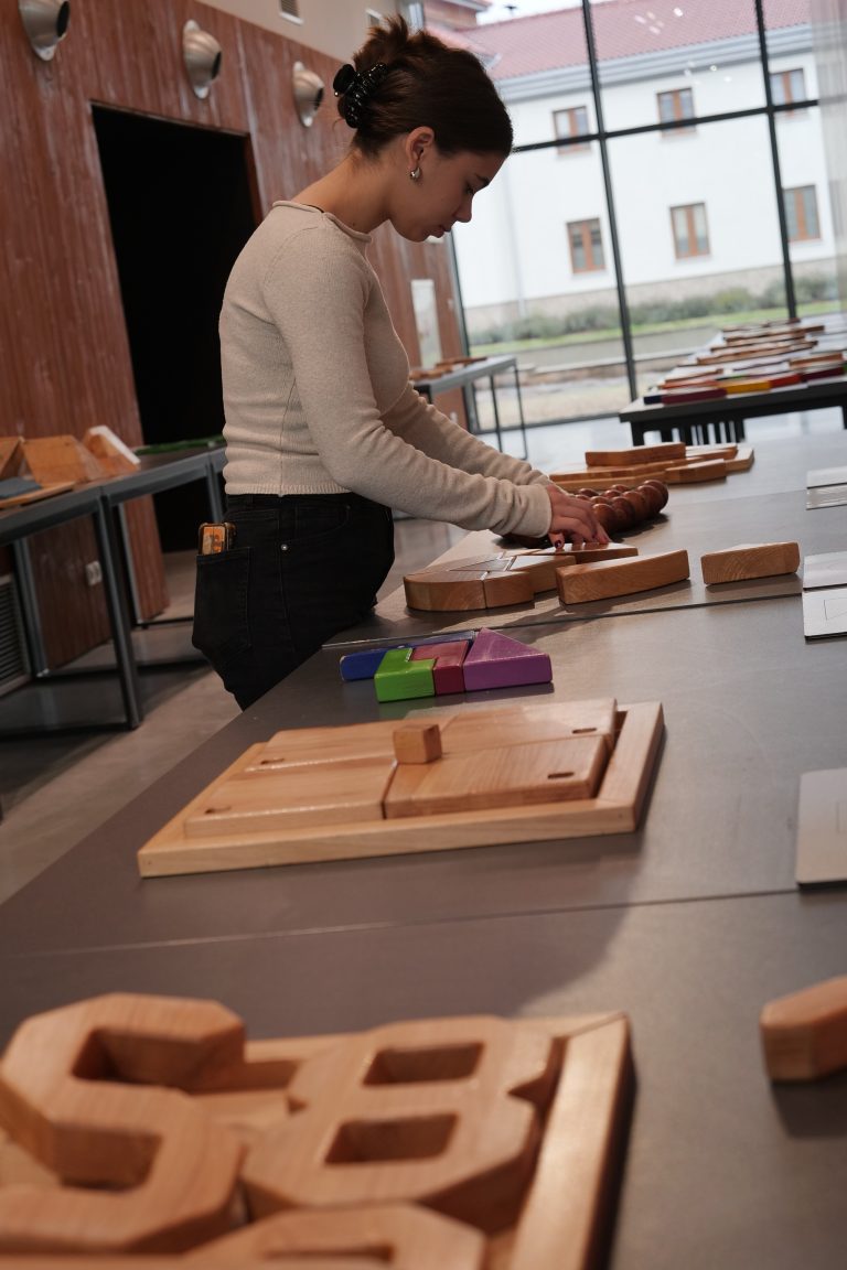 Młoda kobieta w jasnym swetrze wchodzi w interakcję z drewnianym eksponatem edukacyjnym, z tyłu widać duże okna z widokiem na zewnątrz.