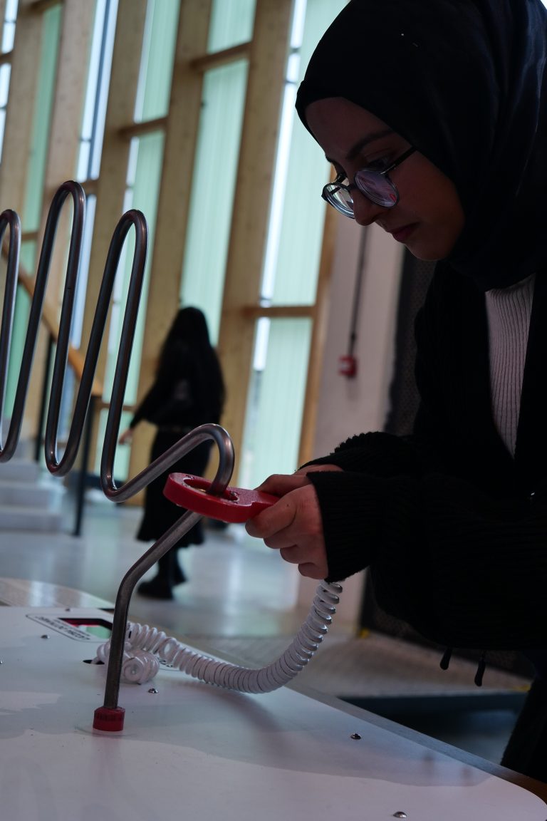 Młoda kobieta w nakryciu głowy i okularach koncentruje się na próbie przeprowadzenia czerwonego kółka przez metalową sprężynę, co jest popularnym eksponatem demonstrującym zręczność i fizykę.