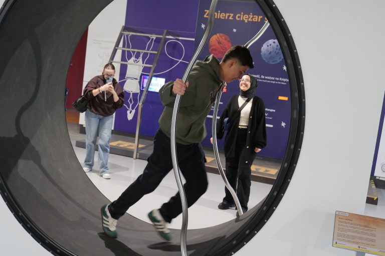 Młody mężczyzna w zielonej bluzie biegnie w dużym, obrotowym kole, podczas gdy dwie inne osoby patrzą na niego, w tle widać tablicę o tematyce kosmicznej.