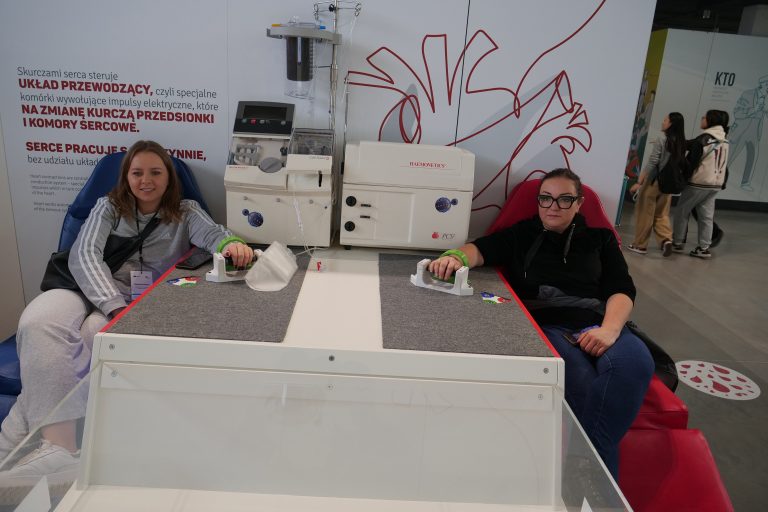 Dwie kobiety siedzą przy interaktywnym eksponacie w muzeum nauki, który symuluje system przewodzący serca, trzymając uchwyty przypominające elektrody.