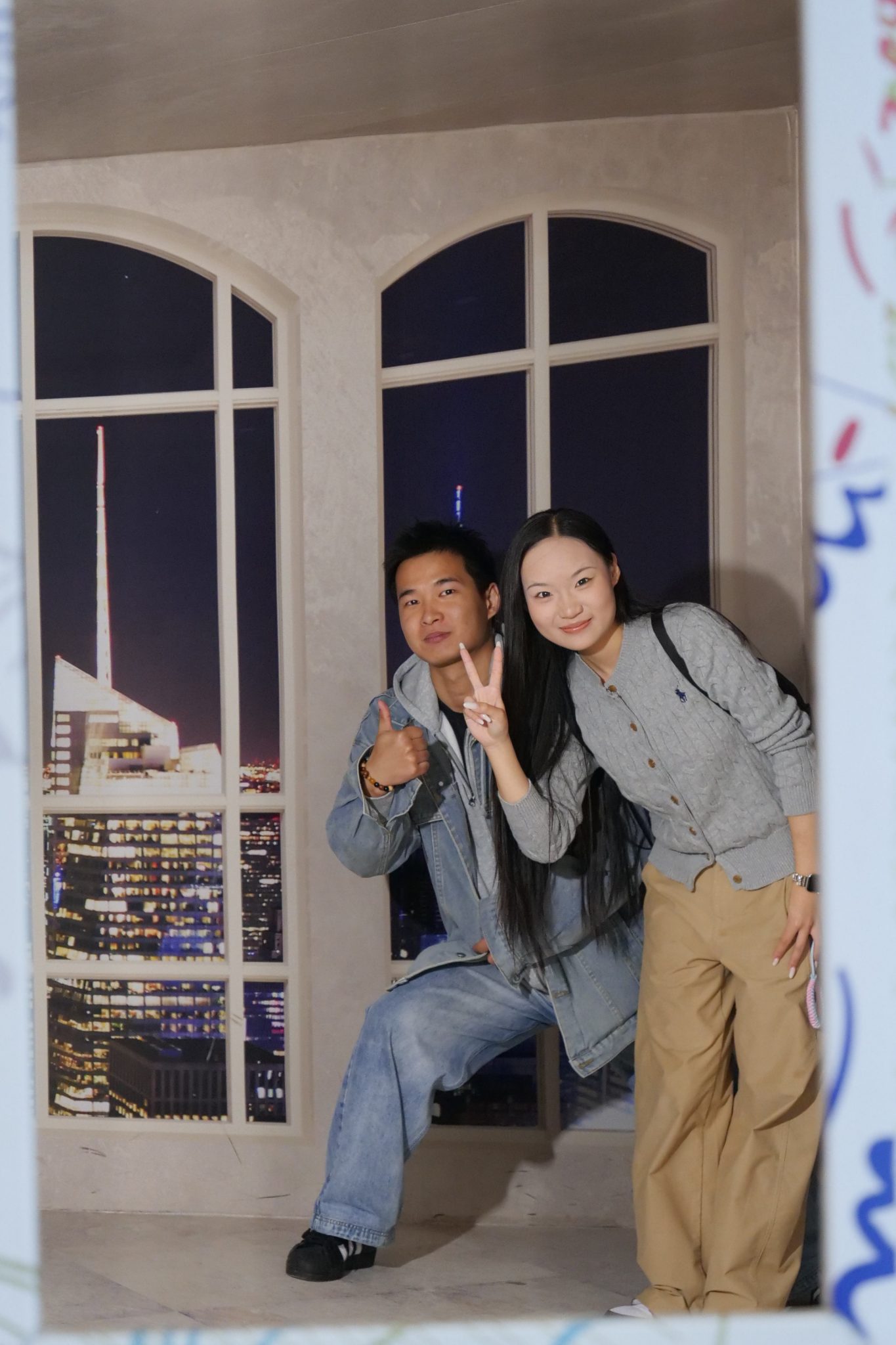 Para młodych ludzi pozuje do zdjęcia w ramce z iluzją optyczną, naśladującą wnętrze z dużymi oknami z widokiem na oświetlone miasto nocą.