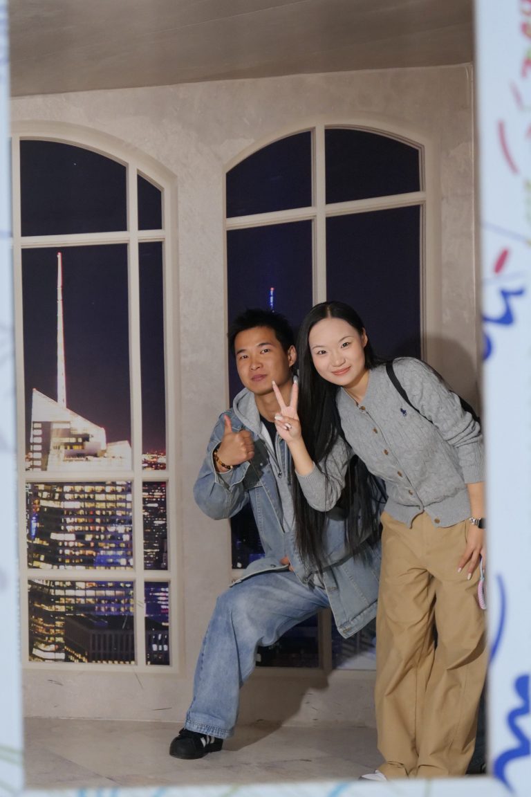 Para młodych ludzi pozuje do zdjęcia w ramce z iluzją optyczną, naśladującą wnętrze z dużymi oknami z widokiem na oświetlone miasto nocą.