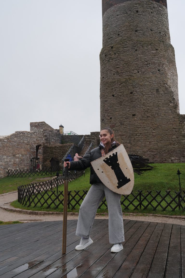 Studentka wymiany międzynarodowej z mieczem i tarczą na tle Zamku Królewskiego w Chęcinach.