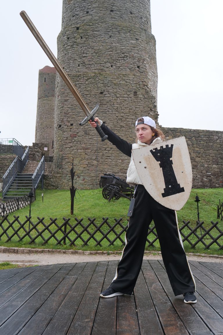 Studentka wymiany międzynarodowej z mieczem i tarczą na tle Zamku Królewskiego w Chęcinach.