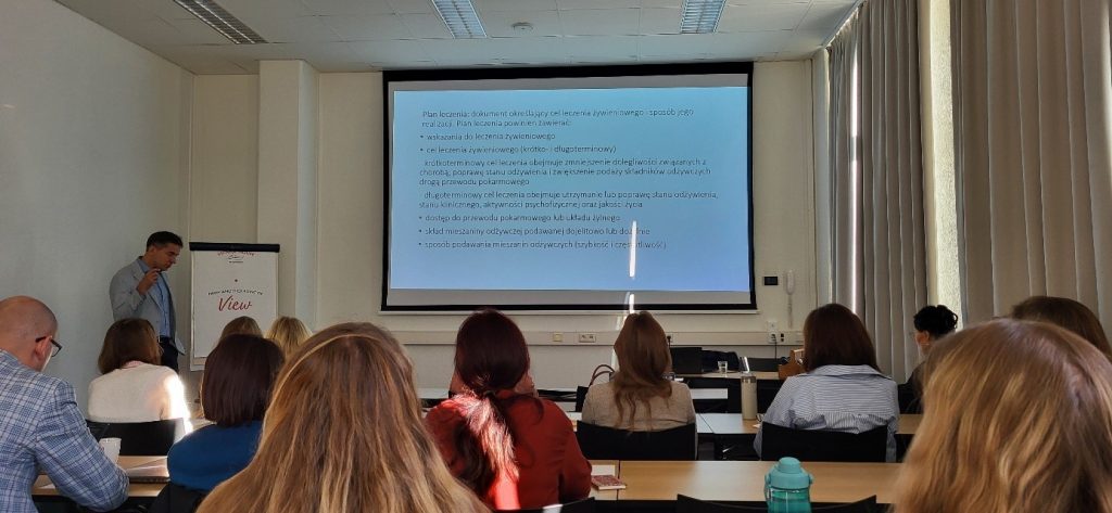 Uczestnicy kursu siedzą w sali ćwiczeniowej, patrzą na ekran z wyświetloną prezentacją.