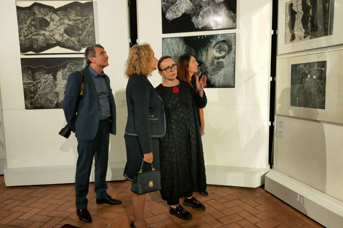 Cztery osoby stoją w galerii sztuki, wpatrując się w wiszące na ścianie czarno-białe grafiki o abstrakcyjnych, organicznych kształtach. Kobieta w centrum, w czarnej sukience i okularach, unosi rękę w geście tłumaczenia czegoś pozostałym. Obok niej stoi kobieta w granatowym kostiumie oraz mężczyzna w garniturze z przewieszonym aparatem fotograficznym.