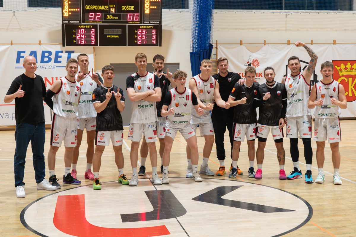 Grupa koszykarzy i trenerów stoi na środku hali sportowej. Wszyscy uśmiechają się i pokazują gesty radości po wygranym meczu. W tle widać tablicę wyników oraz banery sponsorów.