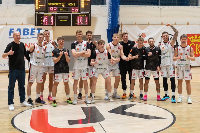 Grupa koszykarzy i trenerów stoi na środku hali sportowej. Wszyscy uśmiechają się i pokazują gesty radości po wygranym meczu. W tle widać tablicę wyników oraz banery sponsorów.