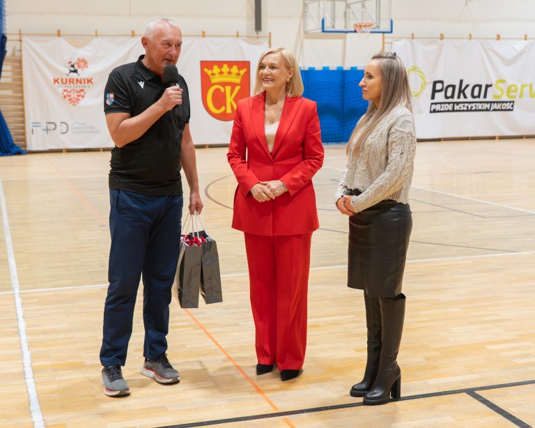 Na boisku stoją trzy osoby. Mężczyzna trzyma mikrofon i torbę z upominkami, obok niego stoją dwie kobiety. Wszyscy uśmiechają się podczas krótkiej ceremonii. W tle banery i sprzęt sportowy.