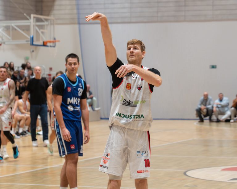 005.jpg: Koszykarz w białym stroju z numerem 10 jest w trakcie wykonywania rzutu wolnego. Jego prawa ręka jest wyciągnięta w górę w geście rzutu. Obok stoi zawodnik drużyny przeciwnej w niebieskim stroju z napisem "MKS".