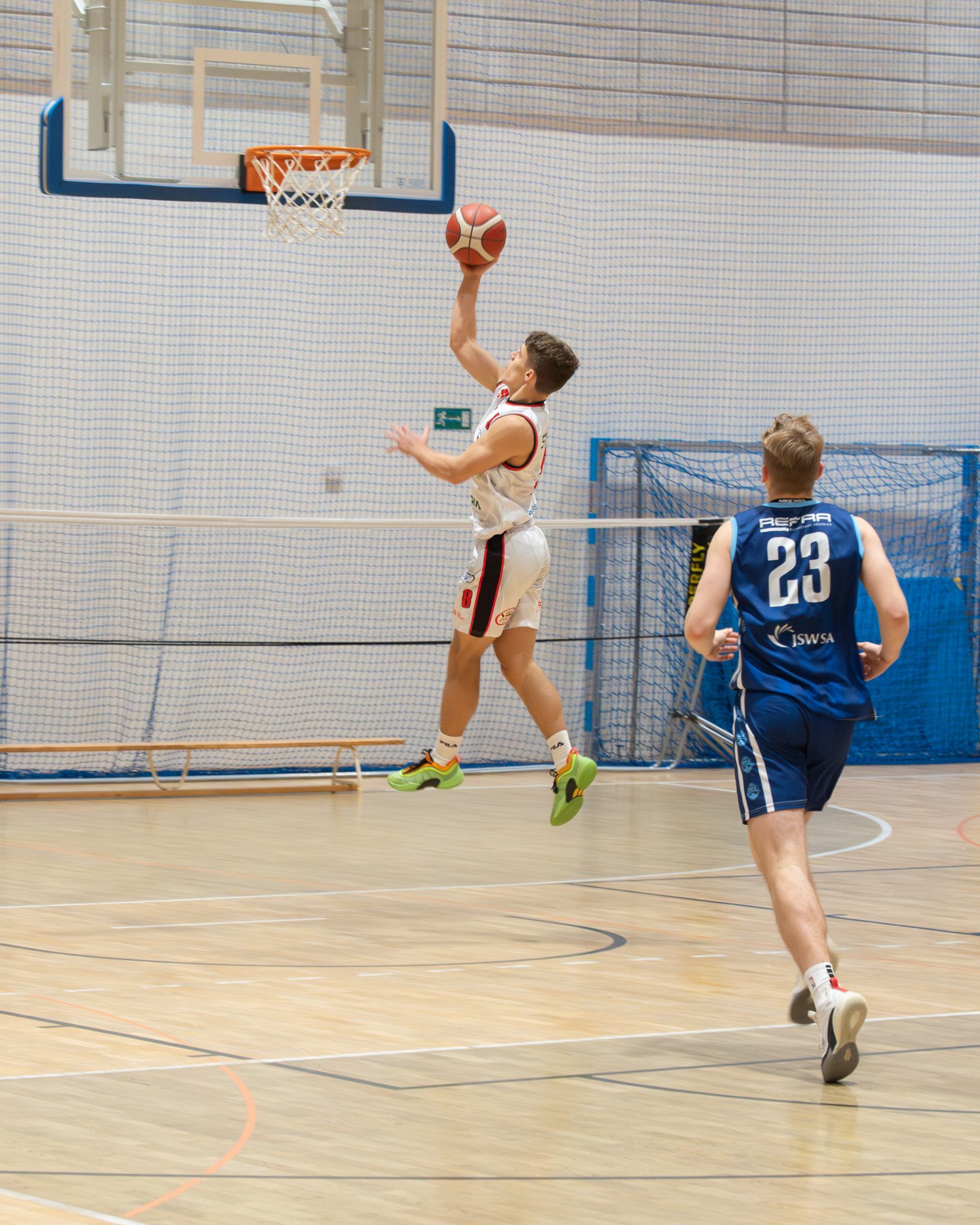 Dynamiczne ujęcie koszykarza w białym stroju, który jest w powietrzu i wykonuje rzut (layup) prawą ręką do kosza. Z tyłu próbuje go dogonić obrońca w niebieskim stroju z numerem 23, aby zablokować rzut.