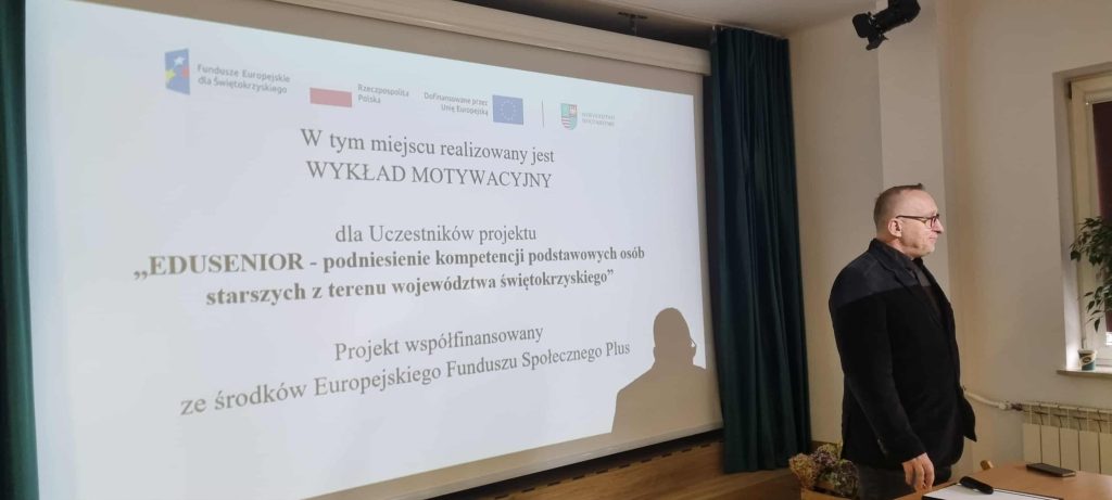 Prowadzący wykład stoi przed ekranem, na której widać planszę z nazwą projektu oraz tytuł wykładu.
