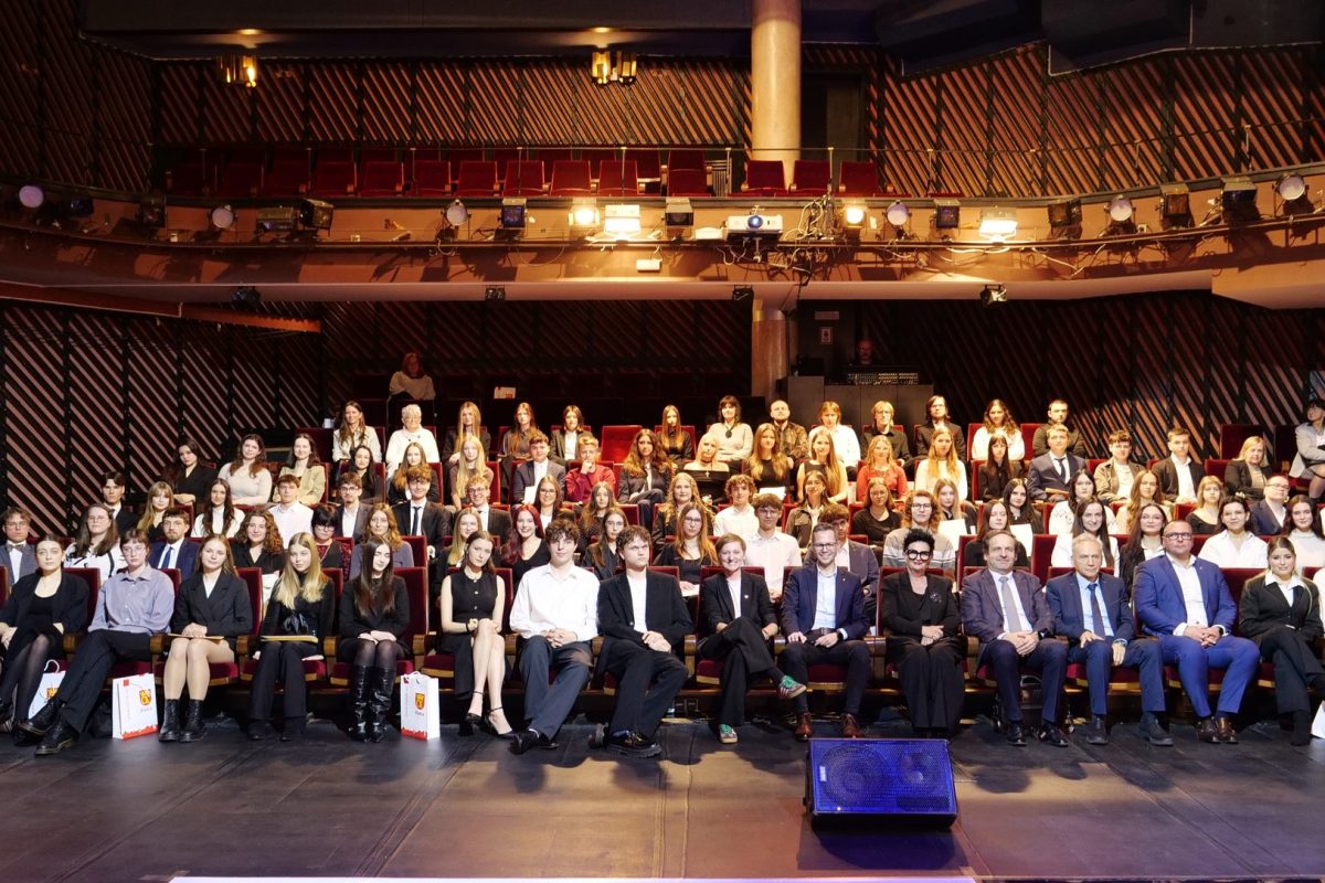 Grupa kilkudziesięciu osób siedzi w kilku rzędach eleganckiej sali teatralnej. W pierwszym rzędzie są dorośli i młodzież w formalnych strojach, obok części leżą torby upominkowe. Za nimi siedzi liczna, odświętnie ubrana młodzież. W tle widoczne są rzędy bordowych foteli, balkon i oświetlenie sceniczne.