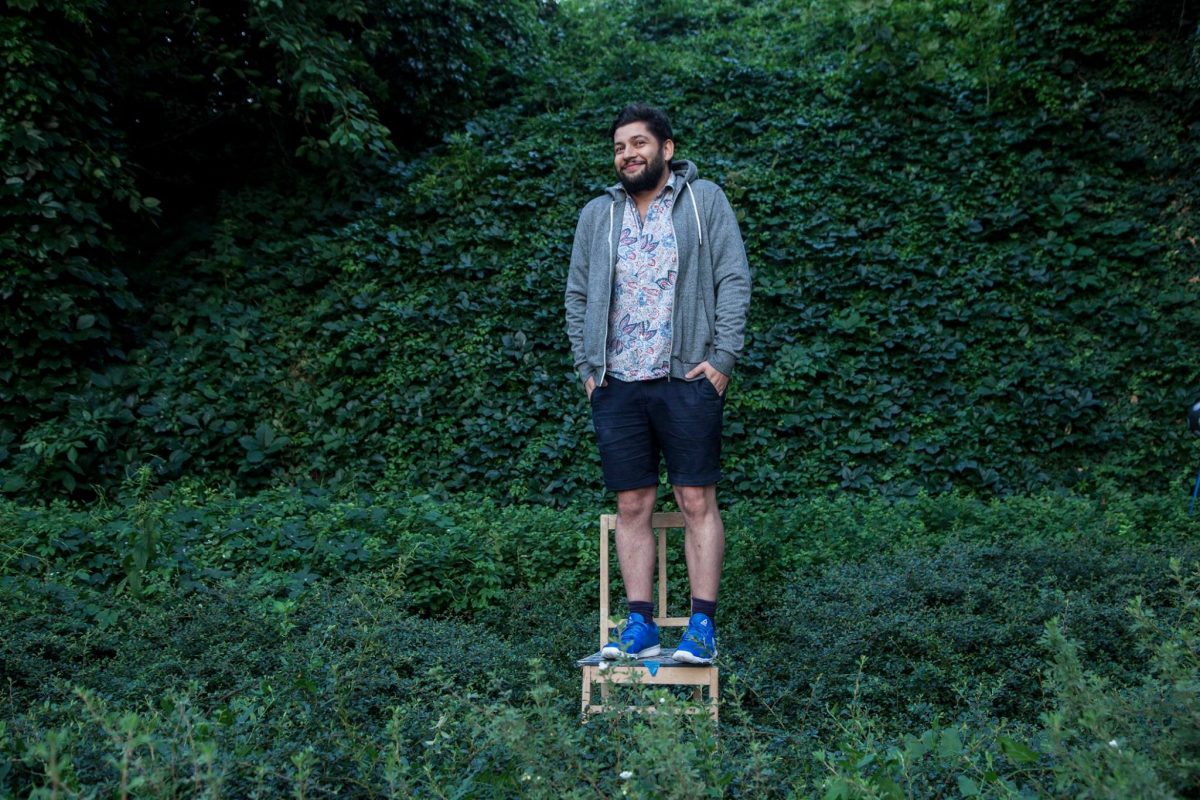 Mężczyzna z brodą stoi na drewnianym krześle na tle gęstej ściany zielonych liści i bluszczu. Ma na sobie wzorzystą koszulę, szarą rozpinaną bluzę, ciemne szorty i jaskrawoniebieskie buty sportowe. Patrzy w bok z lekkim uśmiechem, trzymając ręce w kieszeniach.